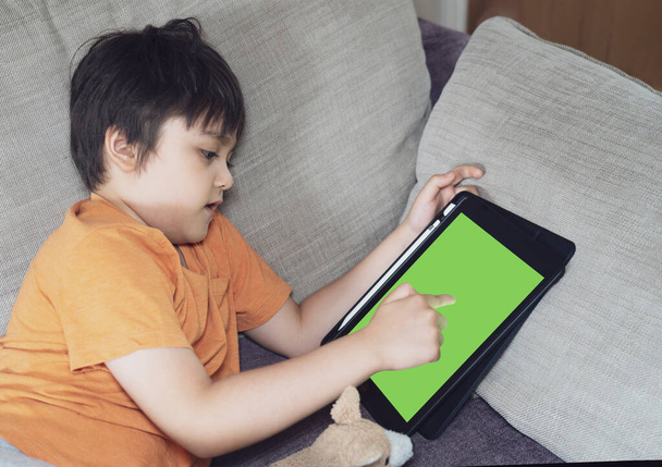 子供はタブレット上でソファ遊びゲームに横たわって、子供の男の子は自宅でリラックスしたり、デジタルパッドで書いたり、新しい通常の生活教育や距離学習の概念のための子供の活力 - 写真・画像