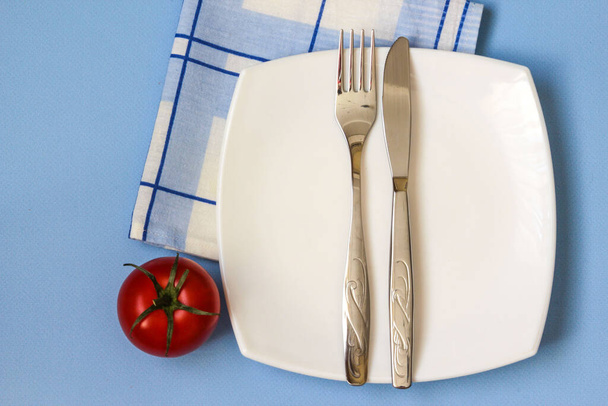 Fork, knife and white plate with tomate over blue background. Clean plate and cutlery on light background. Top view. - Valokuva, kuva