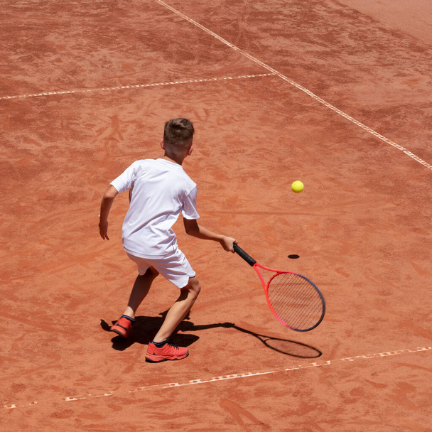 Boy plays tennis on a clay tennis court. Child is concentrated and focused on the game. Young tennis player with racket in action. Kids tennis sport concept. Motion. Shadow. Copy space - Photo, Image