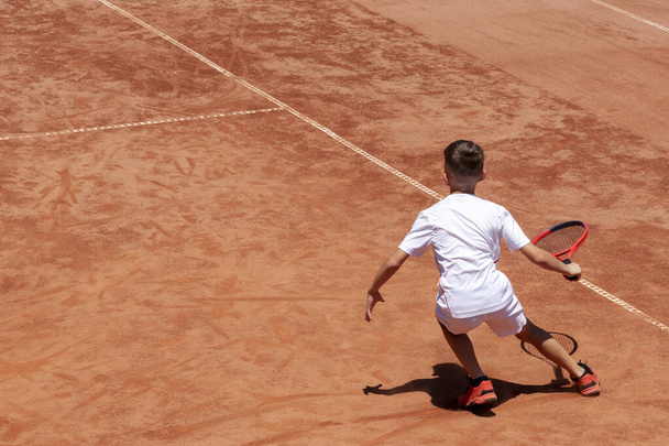 Jonge mannelijke tennisspeler met racket in actie. Jongen speelt tennis op een tennisbaan. Kind is geconcentreerd en gericht op het spel. Kids tennis sport achtergrond met schaduw. Beweging. Kopieerruimte - Foto, afbeelding