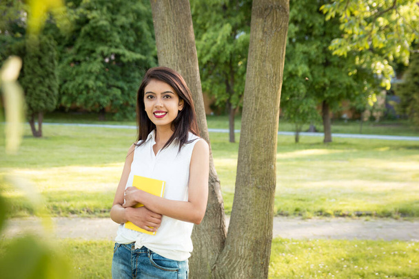 Joven chica india hermosa de pie en el parque cerca de un árbol, sosteniendo un cuaderno amarillo en sus manos. Tiempo libre del estudio o del trabajo
. - Foto, imagen