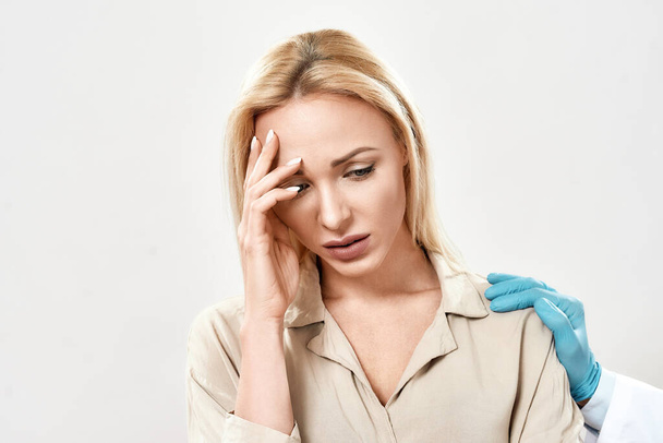 Portrait of depressed young woman visiting doctor. Doctor in blue sterile gloves supporting and comforting upset female patient standing against grey background in studio - 写真・画像