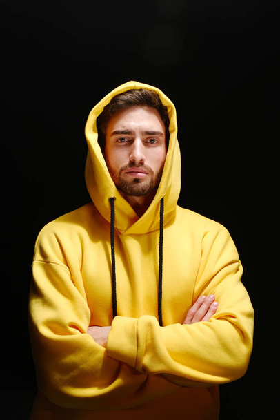 A young man of 25-30 years old in a yellow sweatshirt stands against a black wall.  - Photo, Image