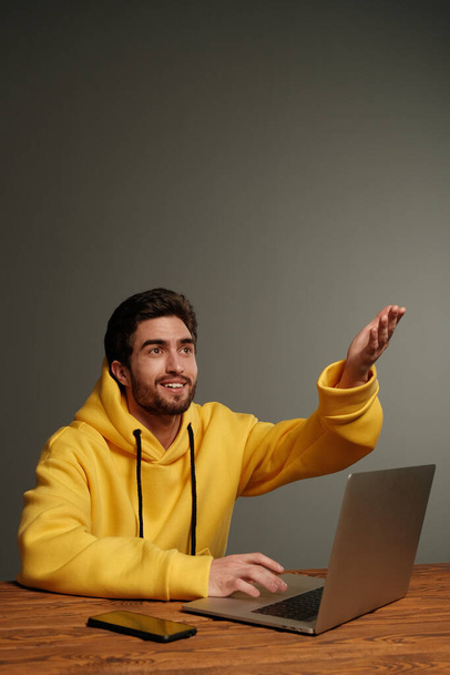 A young guy with different emotions in a yellow sweatshirt works using a laptop and smartphone.  - Photo, Image