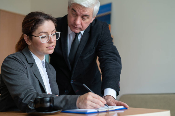 The male senior director watches as the female secretary signs the papers. Business woman fills out documents under the supervision of a boss in the office. - Valokuva, kuva
