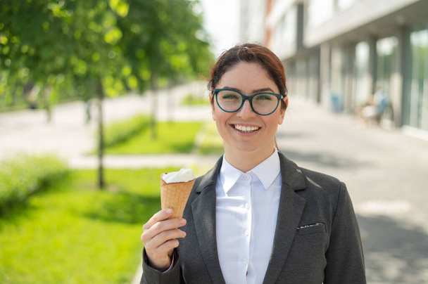 businesswoman is walking outdoors and eating vanilla icecream on a sunny summer day. Portrait of a smiling girl in a white shirt and a jacket with an ice cream cone. Lunch break of an office employee. - Foto, Bild