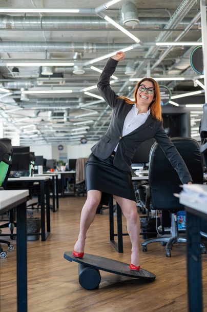 Cheerful business woman in red high heel shoes lost equilibrium on the balance board and falls. A female employee in a suit does sports at the workplace during the break. Bad coordination. - Foto, Imagem