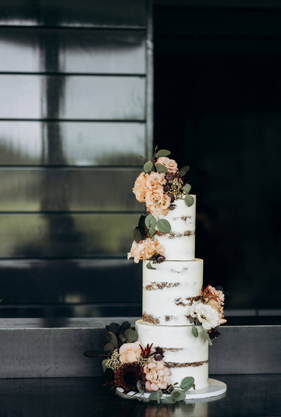 torta de boda con gradas blancas decoradas con hojas de eucalipto, rosas rosadas en polvo y claveles
 - Foto, Imagen