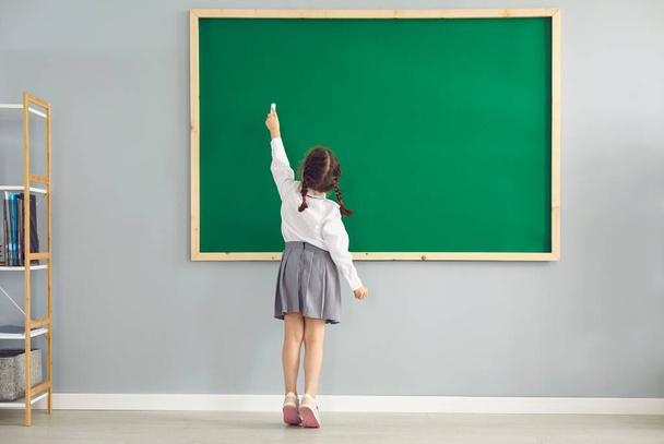 Back view of little girl writing on blackboard with chalk at classroom. Smart first grader answering on lesson at school - 写真・画像