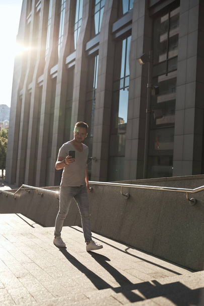 a stylish guy, wearing glasses, climbs the steps to the office check-in with a phone in his hands. light jeans and a fashionable gray T-shirt. contour sunlight - Photo, Image
