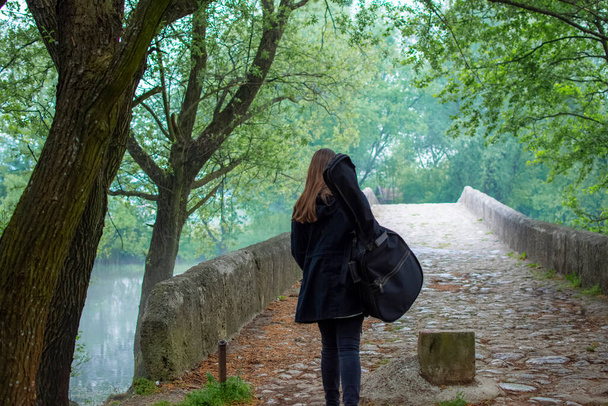 Νεαρή κοπέλα κουβαλάει τσάντα κιθάρας ενώ περπατάει μόνη της στο μαντρί. Γυναικείος κιθαρίστας ταξιδεύει με κιθάρα και απολαμβάνει την όμορφη θέα - Φωτογραφία, εικόνα