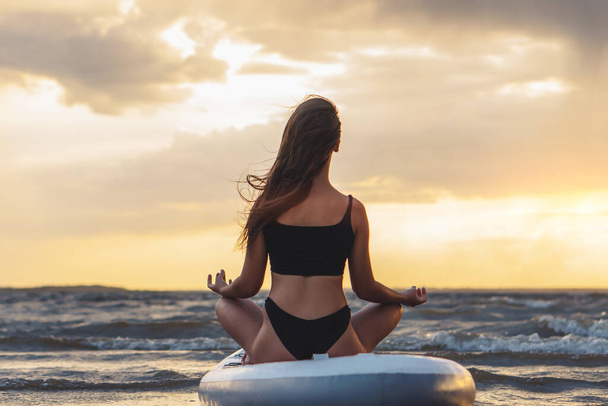 Young sexy woman surfer meditating on her sup board on the sunset. Rest at nature concept - 写真・画像