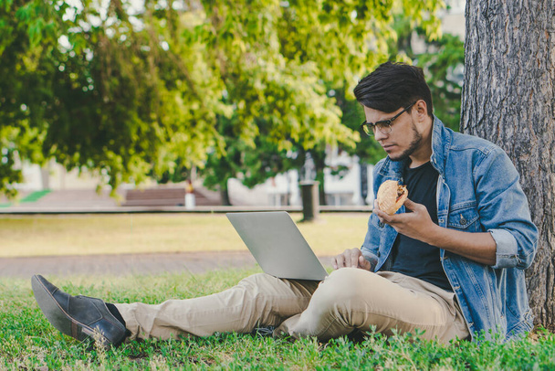 Joven freelancer guapo trabajando en un portátil mientras come al aire libre. Concepto de trabajo exterior
. - Foto, Imagen