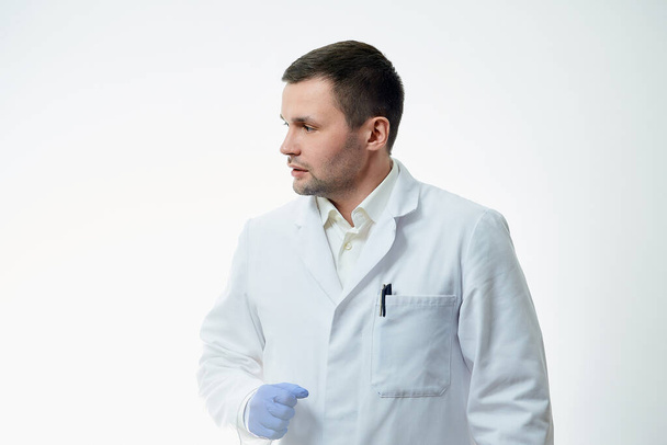 Ένας άντρας καυκάσιος γιατρός με άσπρη ποδιά και μπλε αναλώσιμα ιατρικά γάντια γύρισε το κεφάλι του στο πλάι. Ένας επιστήμονας απομονώνεται σε λευκό φόντο. - Φωτογραφία, εικόνα