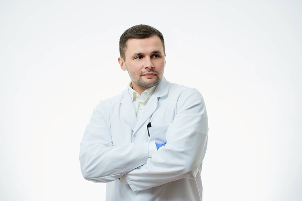 A male caucasian doctor in a white lab coat and blue disposable medical gloves is staying crossed his arms over his chest. A scientist is isolated over white background. - Fotoğraf, Görsel
