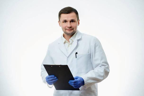 Un medico caucasico maschio gentile con un camice bianco e guanti medici monouso blu sta tenendo un cartone con una scheda paziente con una diagnosi. Uno scienziato è isolato su sfondo bianco. - Foto, immagini