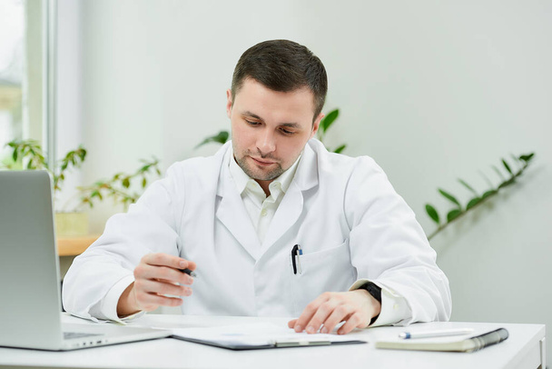 Um médico caucasiano sério em um jaleco branco está sentado em sua mesa e examinando o histórico médico do paciente em um hospital. Um terapeuta perto de um laptop no consultório de um médico
. - Foto, Imagem