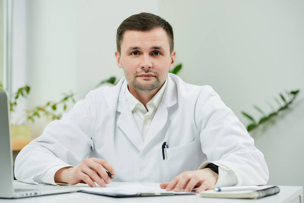 Ένας λευκός γιατρός με άσπρη ποδιά κάθεται στο γραφείο του και αποσπάται από την εξέταση του ιατρικού ιστορικού του ασθενούς σε νοσοκομείο. Ένας θεραπευτής κοντά σε ένα φορητό υπολογιστή στο γραφείο ενός γιατρού.  - Φωτογραφία, εικόνα