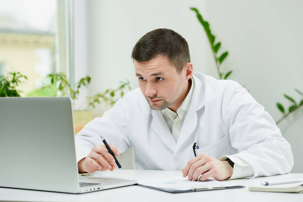 Vážný běloch v bílém laboratorním plášti pečlivě zapisuje pacientovu anamnézu do počítače v nemocnici. Terapeut sedí u stolu s laptopem v ordinaci lékaře. - Fotografie, Obrázek
