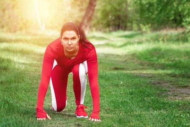 A girl in a tracksuit is preparing to run, physical exercises against the backdrop of nature, prelaunch pose. The concept of a healthy lifestyle, exercise, fresh air. Copy space - Фото, изображение