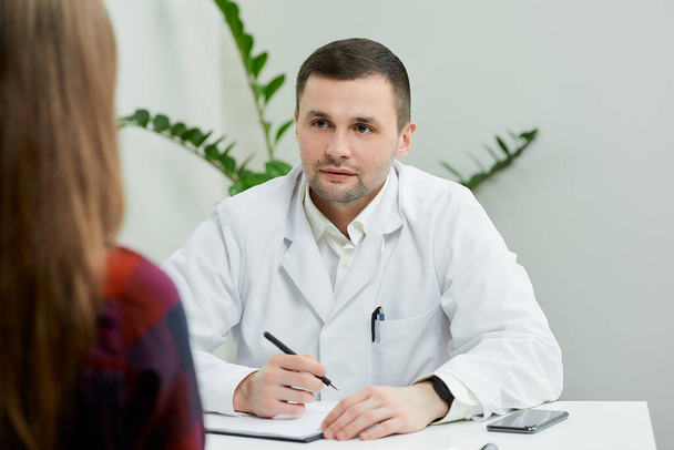 Un ritratto di un medico con un camice bianco seduto alla scrivania che ascolta una paziente nello studio di un medico. Una donna ad un appuntamento dal medico in ospedale. - Foto, immagini