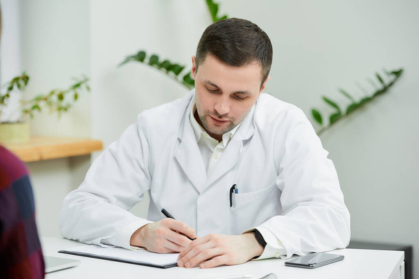 A portrait of a doctor in a white lab coat who is sitting at a desk and writing a diagnosis to a female patient in a doctor's office. A woman at a doctor's appointment in a hospital. - Foto, immagini