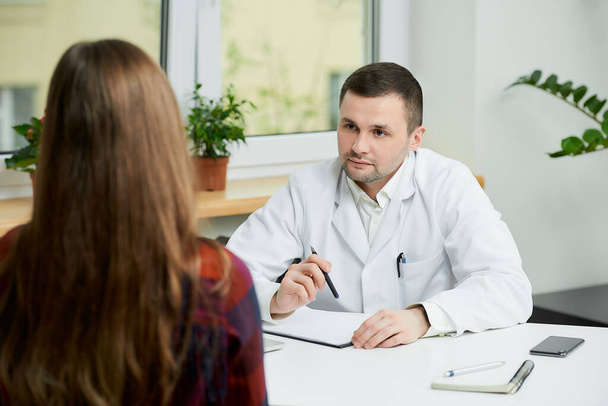 Un médecin caucasien sérieux en blouse blanche est assis à un bureau près d'une patiente dans le bureau d'un médecin. Une femme aux cheveux longs au rendez-vous chez le médecin à l'hôpital. - Photo, image