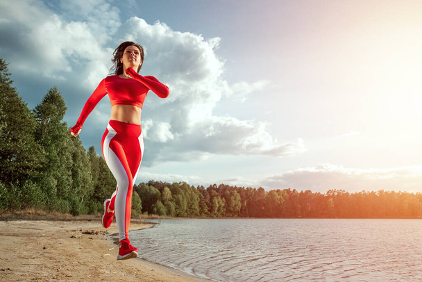 A beautiful, happy girl in a tracksuit runs along the path of a lake or sea. The concept of a healthy lifestyle, cardio workout. Copy space - Valokuva, kuva