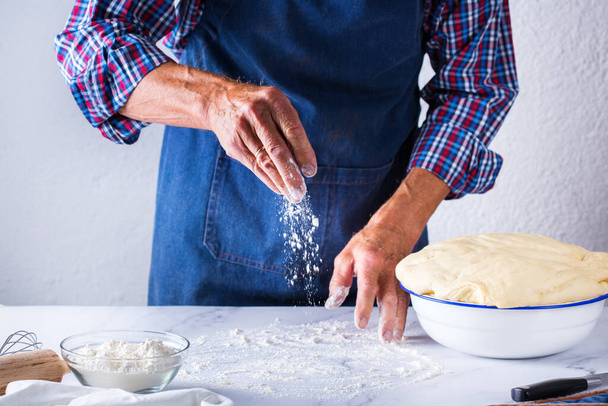 Backen, zu Hause essen, gesunde Ernährung und Lifestylekonzept. Senior Bäcker Mann Kochen, Kneten frischen Teig mit den Händen, Rollen mit Nadel, Ausbreiten der Füllung auf der Torte auf einem Küchentisch mit Mehl - Foto, Bild
