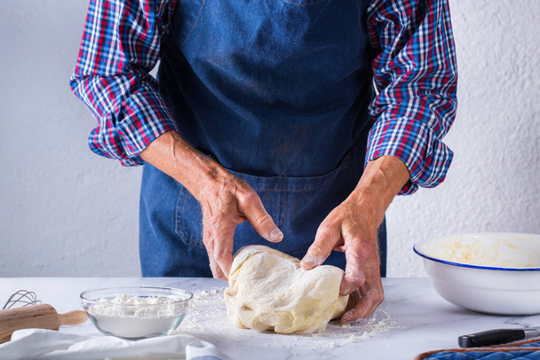 Baking, eating at home, healthy food and lifestyle concept. Senior baker man cooking, kneading fresh dough with hands, rolling with pin, spreading the filling on the pie on a kitchen table with flour - Photo, Image