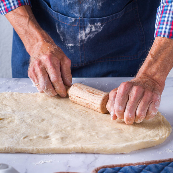 Baking, eating at home, healthy food and lifestyle concept. Senior baker man cooking, kneading fresh dough with hands, rolling with pin, spreading the filling on the pie on a kitchen table with flour - Photo, image