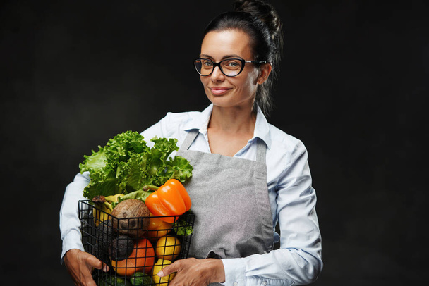 エプロンの中年女農家の肖像は、新鮮な野菜や果物のバスケットを保持しています - 写真・画像