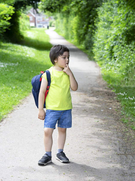 Kid with standing alone and puting finger on his mouth with bored face, Unhappy kid get bored waiting for school bus, School boy carrying backpack get ready for school summer camp - Foto, afbeelding