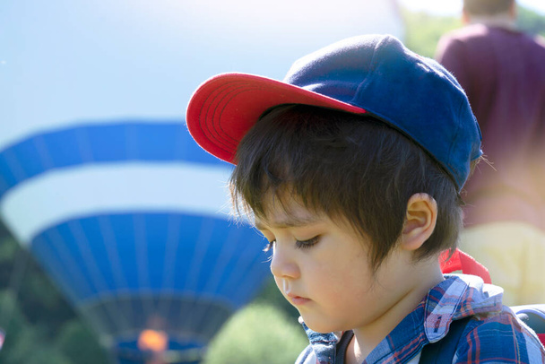 Egy fiú ül egyedül a parkban egy forró napsütéses napon, unatkozó arccal, boldogtalan gyerek ül a füvön kék sapkában, arra várva, hogy lássa a hőlégballont felemelkedni az égbe, szabadtéri tevékenységek - Fotó, kép