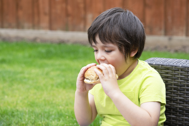 Cute happy preschool kid eating hamburger sitting in the garden. Healthy child eating delicious homemade burger outdoors on summer day - Photo, Image