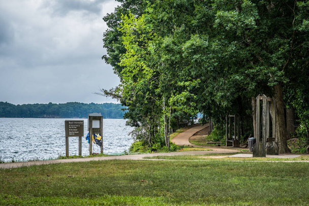 Ścieżka chodnikowa nad jeziorem biegnie na plażę przy kamizelkach ratunkowych z ławkami huśtawkowymi do oglądania jeziora, podczas gdy reszta szlaku biegnie do lasów wzdłuż brzegu - Zdjęcie, obraz