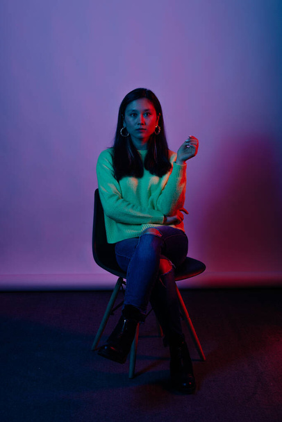 Vertikales Porträt einer jungen Frau, die auf einem Stuhl im Studio sitzt, mit rotem und blauem Licht auf weißem Hintergrund. - Foto, Bild