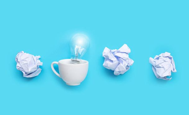 Glühbirne in weißer Tasse mit weißen zerknüllten Papierkugeln auf blauem Hintergrund. Ideen und kreatives Denken. Kopierraum - Foto, Bild