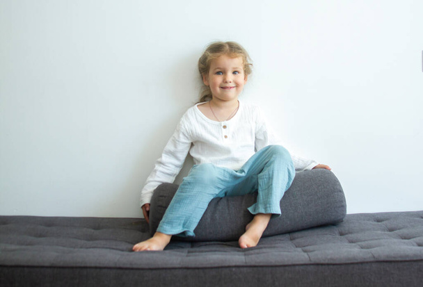 Little cute girl in a bright room, a minimum of objects in the room, the child rejoices, sitting on a gray sofa against a white wall. - Photo, Image