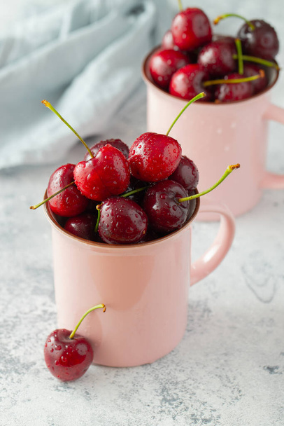Dos tazas rosadas con cerezas frescas maduras. Bayas orgánicas dulces sobre un fondo de hormigón ligero
 - Foto, imagen