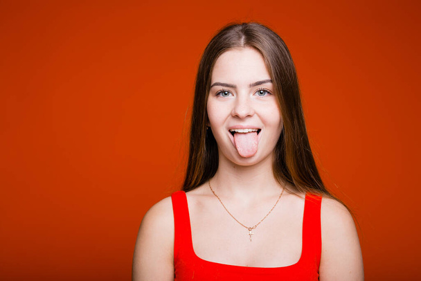 Cute naughty girl of European appearance shows her tongue to the camera on an orange background - 写真・画像