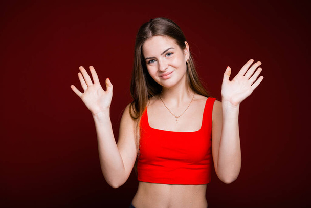 A young European girl with two hands waving and smiling in greeting against a red background - Foto, afbeelding