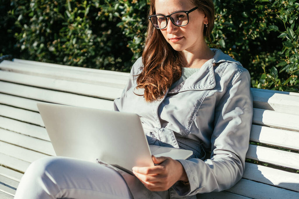 Ragazza freelance caucasica seduta in un parco su una panchina e che lavora su un computer portatile in estate. - Foto, immagini