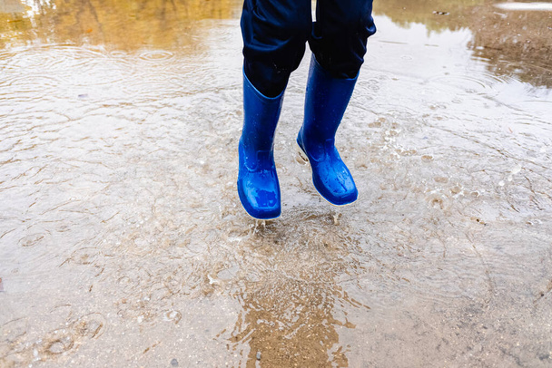 Young boy with blue rubber boots jumps over a puddle of water in a park in his city. - Fotó, kép