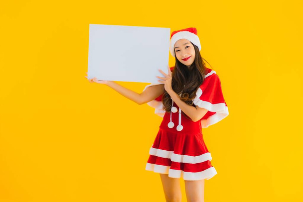 Portret mooie jonge aziatische vrouw kerst kleding en hoed glimlach met lege witte boord teken op geel geïsoleerde achtergrond - Foto, afbeelding