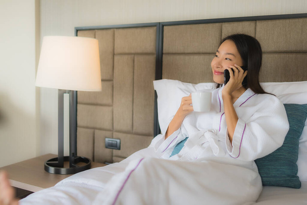 寝室のインテリアでベッドの上にコーヒーカップと携帯電話を使用して肖像画の美しい若いアジアの女性 - 写真・画像