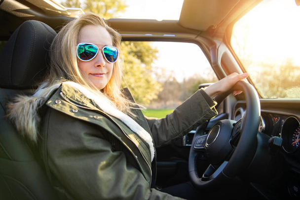 Blond vrouwelijk coureur, rijdend in de natuur in convertible met aviator zonnebril en jas, tijdens zonsondergang - Foto, afbeelding