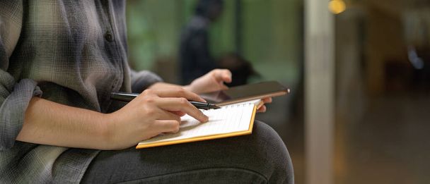 Vista lateral de la trabajadora sentada en la mesa de trabajo y tomando nota en el libro de horarios en blanco mientras mira en el teléfono inteligente
  - Foto, Imagen