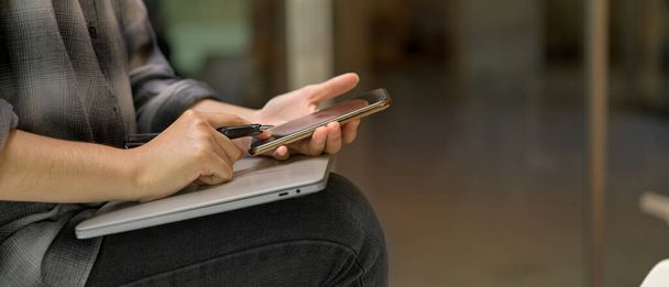 Boční pohled na pracovnici sedící na stole a používající smartphone na notebooku na klíně v pohodlné kancelářské místnosti  - Fotografie, Obrázek