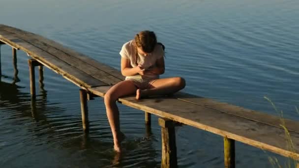 Una bambina con uno smartphone si siede su un molo di legno sul lago durante il tramonto. Attività ricreative all'aperto in estate. Il concetto di un'infanzia felice. - Filmati, video